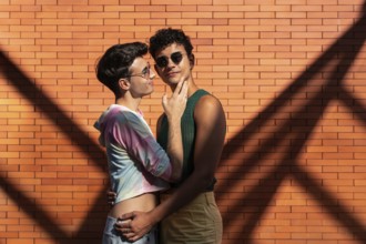A gay couple shares an affectionate embrace against a sunlit brick wall, casting interesting