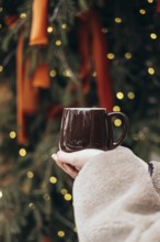Cropped unrecognizable young woman holding a dark brown mug in front of a decorated Christmas tree