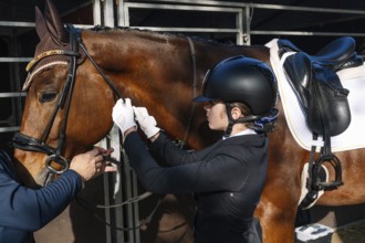 A teenager in classical dressage attire adjusts a horse's bridle with assistance, focusing intently