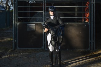 A teenage girl in classical dressage attire confidently holds a saddle. She stands by a stable,