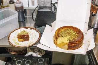 A rich cheesecake topped with crushed pistachios sits in an open box, with a slice served on a