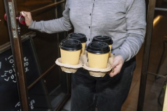 A person opens a cafe door while holding a tray of four takeaway coffee cups. The setting suggests