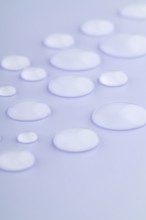 Close-up image of droplets scattered on a smooth, lavender-colored surface The droplets vary in