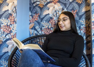 A young woman enjoys reading a book, seated in a modern chair against a colorful floral-patterned
