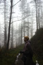 A serene scene of a person in a hoodie walking along a forest trail. Tall trees and fog create a