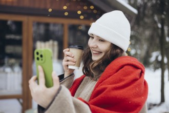 A cheerful young woman in a white beanie and red scarf smiles while holding a coffee cup and taking