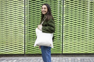 A smiling woman in a green jacket stands in front of a textured green wall, holding a white tote