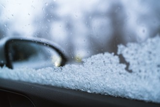 Close-up of a frosted car window with a blurred side mirror visible. The ice crystals and droplets