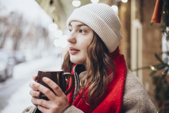 A young woman sips a hot chocolate on a snowy winter day. Dressed warmly in a white beanie and red