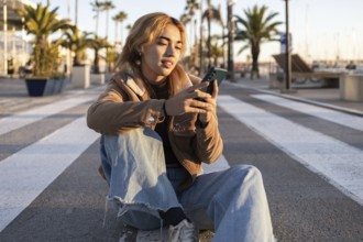 Mixed-race woman sits cross-legged on an outdoor boardwalk, absorbed in her smartphone Palm trees