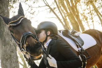 A teenage girl in classical dressage attire gently embraces her horse in a peaceful forest setting,