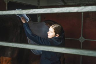 A teenage girl carefully places a saddle blanket on a horse in preparation for a classical dressage