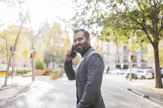 Cheerful Indian man in winter business attire, on a call using a mobile phone on a sunny urban