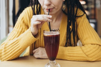 A woman in a yellow sweater sips a refreshing coffee drink through a straw in a cozy cafe setting