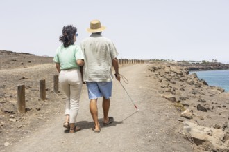 A blind man walking with a female guide along a coastal path. He uses a cane to navigate the rocky