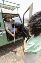 A blind man holds his cane as he steps out of a motorhome, with a woman nearby offering support.