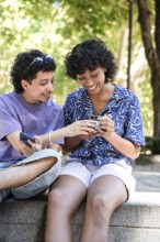A cheerful multiethnic LGBTQ+ couple sitting outdoors, sharing and looking at a phone together,