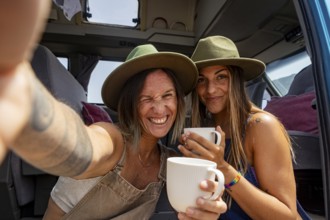 Lesbian couple wearing hats enjoy coffee inside a camper van, capturing a joyful moment of