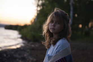 A young girl with wavy hair gazes thoughtfully at the camera while standing by a lakeside at sunset