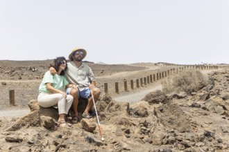 A blind man with a cane and a woman sit together on rocks along a quiet desert path. The scene