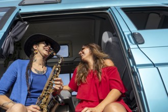 A joyful lesbian couple smiles while relaxing by their van. One plays a saxophone, capturing the
