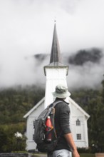 A traveler with a backpack gazes at a quaint white church amid Norway's majestic landscape Misty