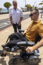 An older man and a younger man in a wheelchair share a moment outdoors on a sunlit pathway by the