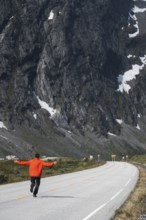 A traveler in a vibrant jacket walks along a winding road, surrounded by Norway's majestic