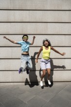 A joyful, multiethnic LGBTQ+ couple, wearing vibrant summer attire, jumps against a concrete wall,