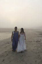 Two women walk along a foggy beach, one in overalls and the other in a flowing white dress The