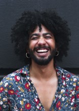 A man with curly hair laughs joyfully, wearing a vibrant floral shirt against a gray background.