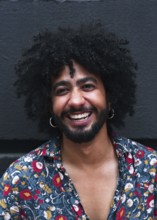 A joyful man with curly hair and hoop earrings smiles against a gray background. Wearing a vibrant
