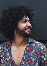 A man with curly hair and a beard smiles while wearing a vibrant floral shirt against a gray