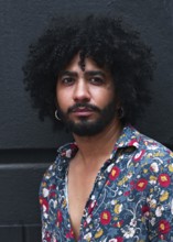 A close up portrait of a man with curly hair, wearing a floral shirt and hoop earrings against a