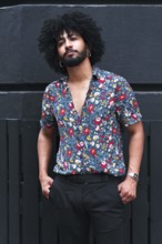 A stylish individual with curly hair and beard leans against a gray wall, wearing a vibrant floral