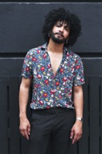 A confident young man with curly hair poses in a vibrant floral shirt against a gray urban wall,