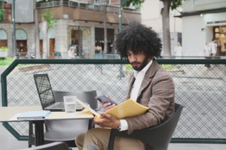 A Mixed-race man with curly hair is engaged in remote work using a laptop and smartphone at an