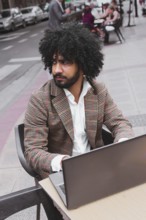 A Mixed-race man with curly hair, dressed in a stylish plaid jacket and white shirt, is focused on