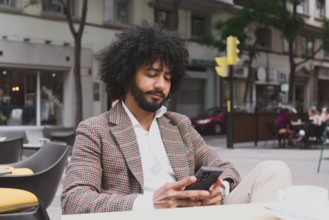 A Mixed-race man with curly hair is engrossed in his smartphone while teleworking on a busy city