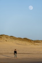 A peaceful scene of a woman standing on the sandy beach at twilight, with a large moon rising above