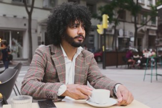 A Mixed-race man with curly hair works remotely, utilizing digital devices at a street cafe while