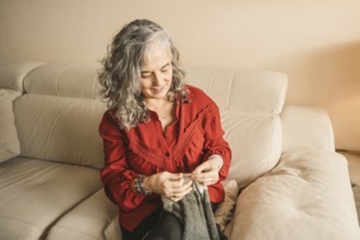 A woman with gray hair sits comfortably on a beige sofa, focused on knitting. She wears a red