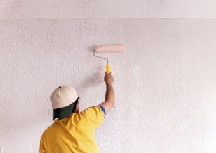 Back view of unrecognizable man in yellow shirt painting a white wall with a roller. This image