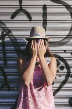 A woman in a straw hat and pink dress stands against a graffiti wall, covering her face with her