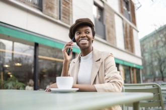 Black woman sits at an outdoor cafe, smiling and talking on her smartphone She is wearing a stylish
