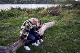 A mother and daughter sit on a wooden log by a scenic riverside, capturing a joyful selfie together