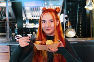 A woman with red hair delights in Asian street food at a lively market She holds chopsticks and a