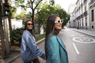 A joyful Latin lesbian couple strolls hand in hand down a tree-lined city street. They wear stylish