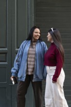 A Latin lesbian couple shares a joyful moment outdoors. One wears a denim jacket and striped shirt,