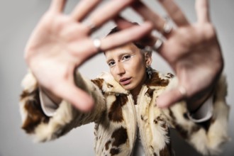 A woman wearing a stylish fur coat poses with hands stretched toward the camera. They sport bold